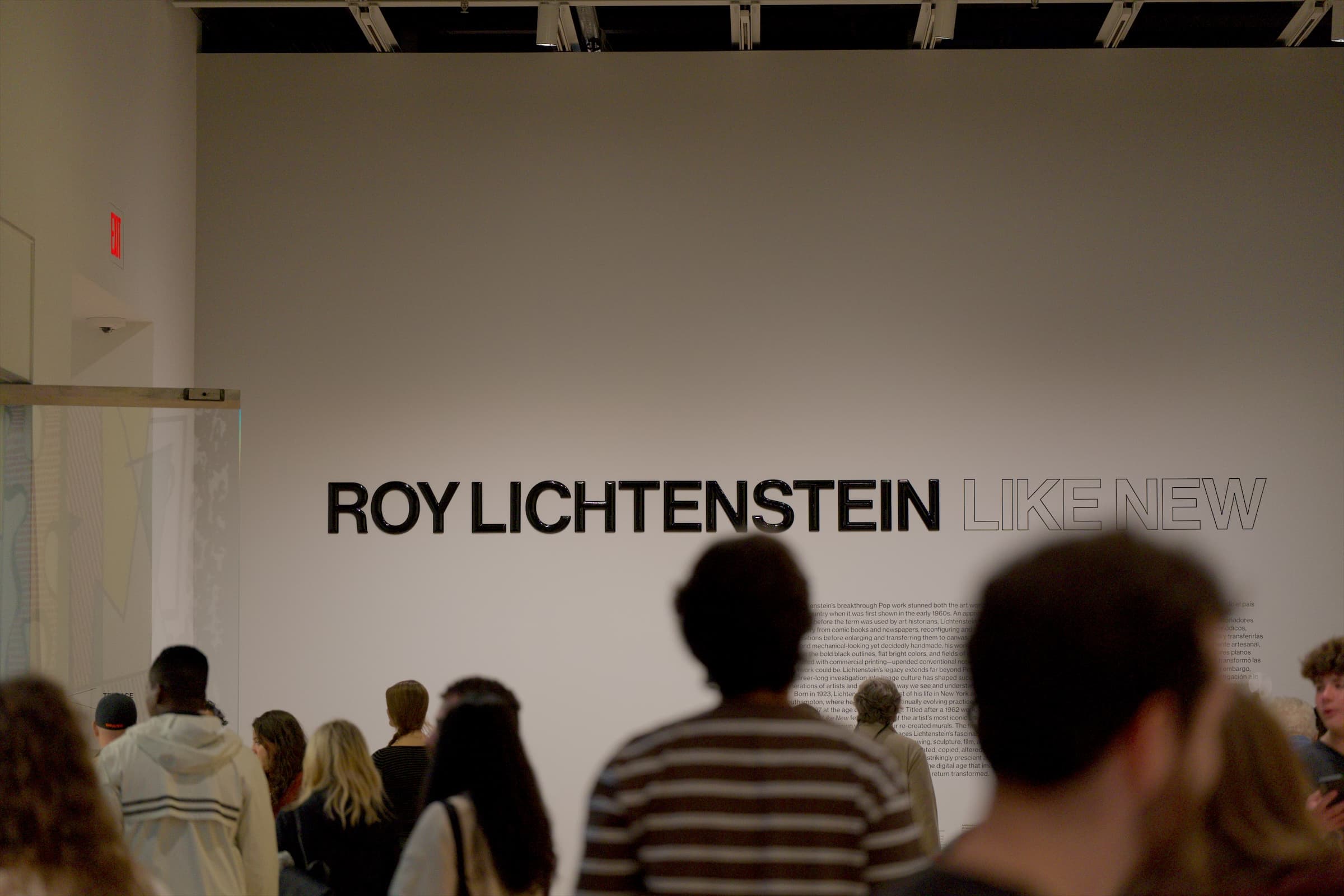A white wall reading ROY LICHTENSTEIN LIKE NEW, with visitors walking past