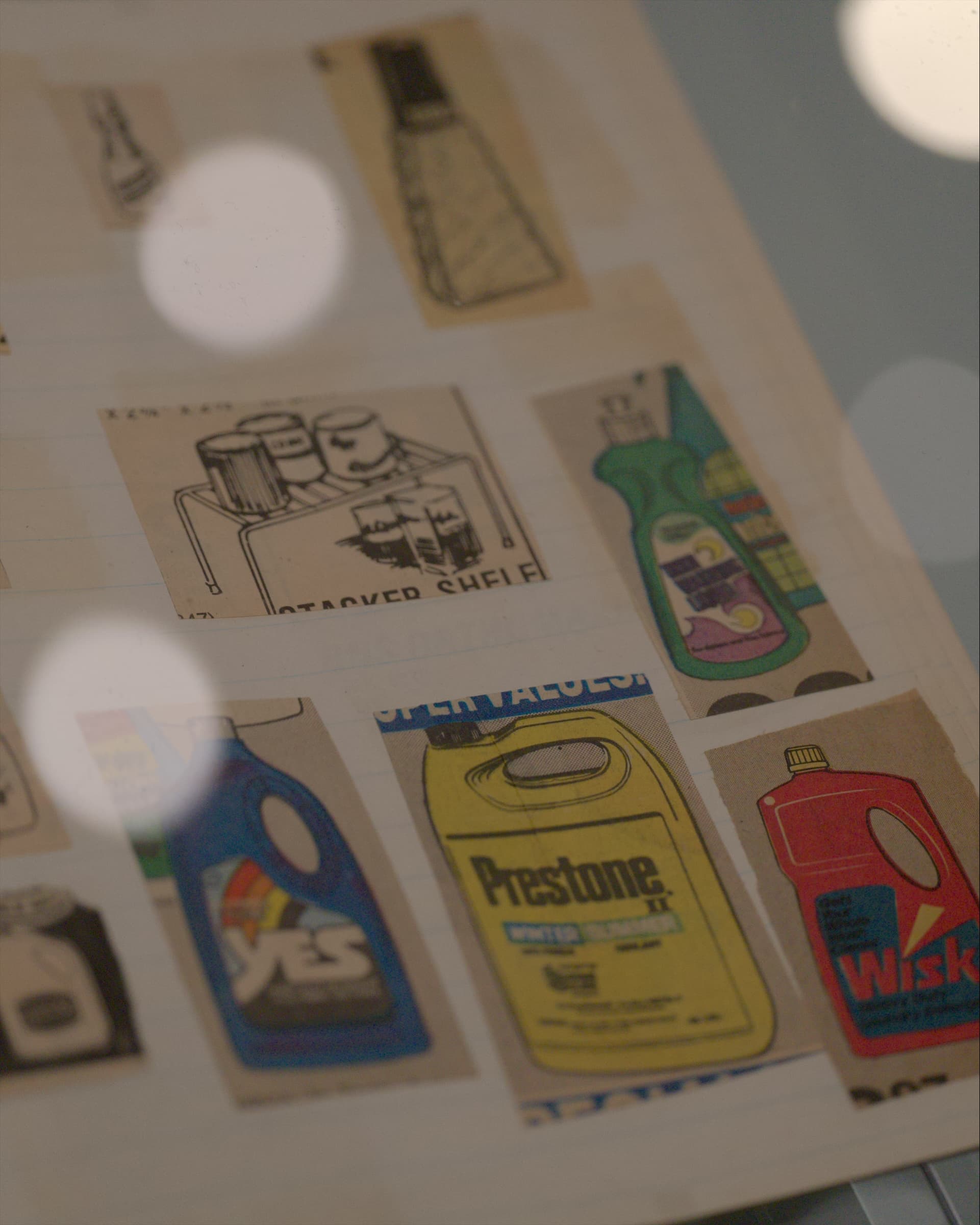 An open sketchbook in a vitrine, pasted with clippings of product ads