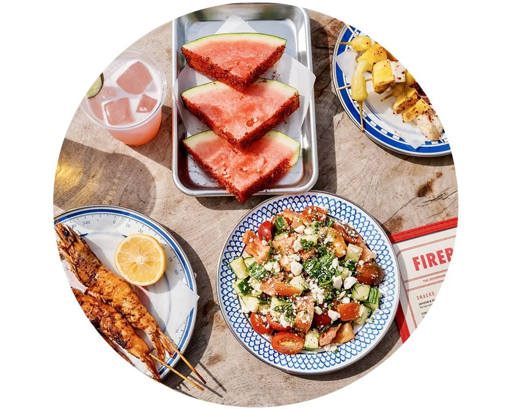 Plates of shrimp skewers, salad and watermelon with a pink cocktail