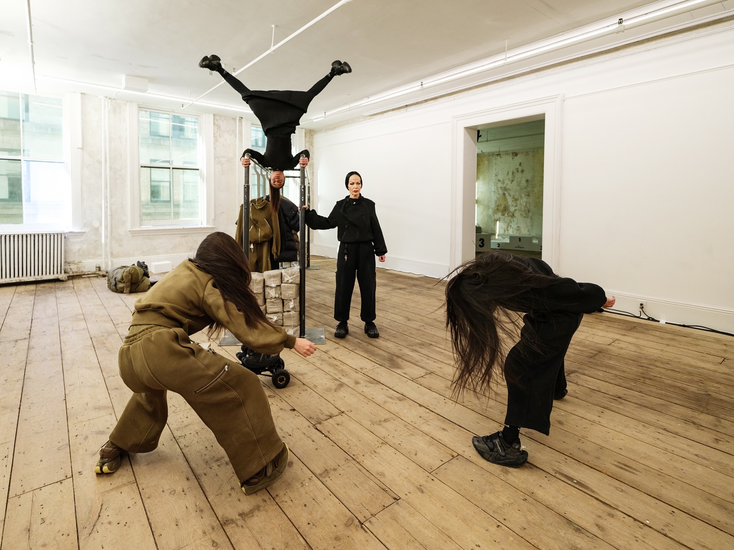 Performers in a white room with an upside-down figure on a rack