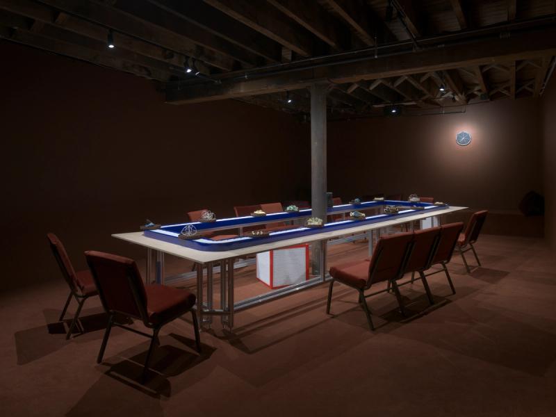 Installation view of the conveyor belt with artworks, surrounded by chairs, in a dark maroon room at Pioneer Works