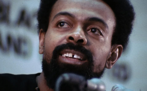 Close-up film still of a bearded man speaking at a microphone